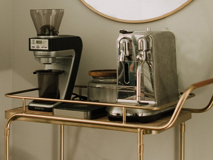Gold metal entertainment cart with coffee grinder machine and silver expresso machine in front of a beige wall. 